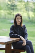 A woman with brown hair sits at a rustic wooden table, surrounded by lush green trees and grass.