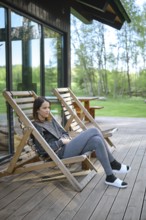 A woman sits comfortably in a wooden deck chair, enjoying the tranquil atmosphere. The lush green