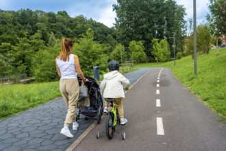 Mother pushing stroller and child riding bicycle on a paved path in a park, enjoying quality family
