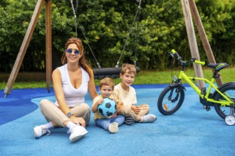 Happy mother sitting with her two sons on a playground's blue rubberized surface near a bicycle and