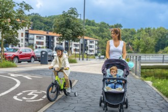 Mother pushing a baby stroller while her son rides a bicycle on a bike path in a modern urban