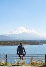 Young man sitting on railings next to a road and looking across the lake to Mt Fuji volcano, Motosu