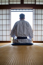Young man wearing kimono sitting in traditional Japanese living room with tatami mats and shoji