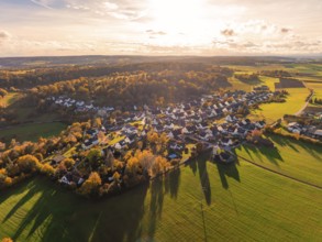 Wide view of a village in autumn atmosphere, surrounded by fields and wooded hills in sunlight,