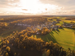 Panoramic autumn view of a village surrounded by golden fields and wooded hills in sunlight,