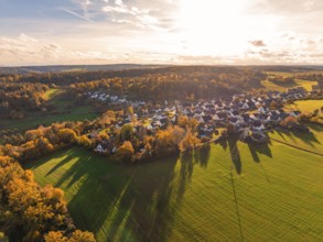 Village in autumn colors with long shadows, surrounded by fields and forests in golden sunlight,