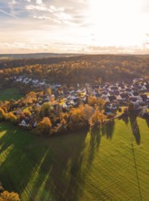 Sun-drenched village with houses and fields in the evening mood, wooded hills in the background,