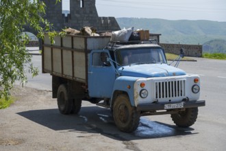 A blue truck transports goods on a rural road with mountains in the background, GAZ-53 loaded with