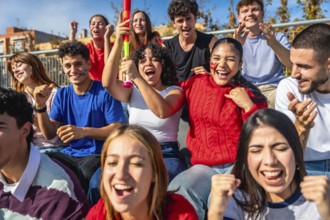 Diverse young friends on stadium bleachers, cheering, clapping and raising hands in daylight as