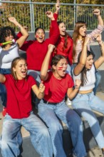 Young women sitting on stadium bleachers, cheering loudly with raised arms and painted faces,