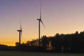 Two wind turbines in front of an orange-purple sky. Trees form a dense silhouette at dusk, Swabian
