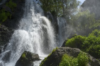 An impressive waterfall falls over rocks surrounded by green vegetation and trees in sunlight,