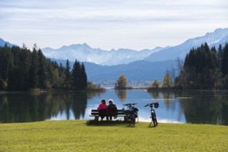Two woman sitting on a bench look at the peaceful Forggensee with mountain views, surrounded by
