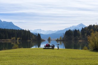Two woman sitting on a bench at Lake Forggensee with a view of mountains, surrounded by autumn
