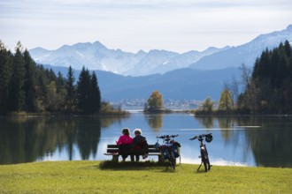 People sitting on a bench with a view over Lake Forggensee to Bergen, surrounded by peaceful nature