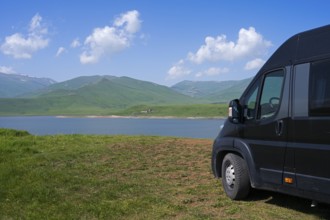 A black van stands at a lake with mountains in the background under a blue sky with clouds, camper,
