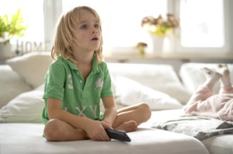 Child, boy, 7 years old, blonde, sitting on sofa, with remote control in hand, watching TV,