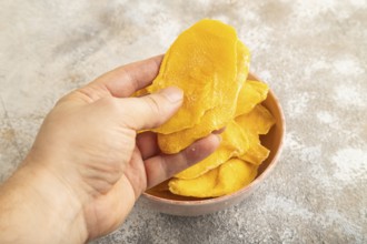 Dried Mango in ceramic bowl with hand on brown concrete background. Side view, close up. healthy