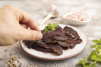 Armenian Basturma dried meat with hand on plate with pepper and herbs on brown concrete background.