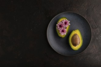 Two Avocado halves with pit and Chrysanthemums on blue plate on black concrete background, top