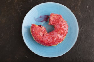 Bitten Pink Donut with sprinkles on blue ceramic plate on black concrete background, top view, flat