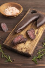 Sujuk sausage on wooden cutting board with pepper and herbs on brown wooden background. Side view,