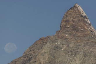 The Matterhorn mountain peak rises steeply against a bright blue sky. The full moon glistens gently