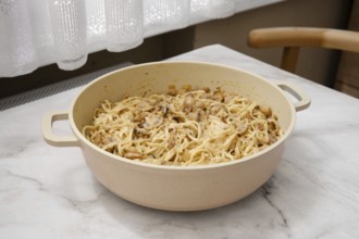 Noodles mixed with mushrooms are being prepared in a large pot placed on a wooden table. The warm