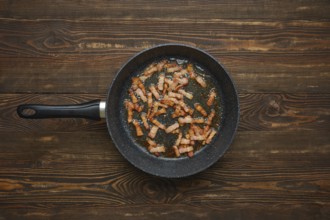 Sizzling strips of bacon fry in a black skillet, releasing savoury aroma and fat on rustic wooden