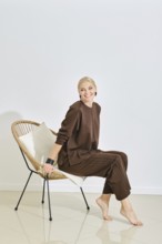 In a modern, bright setting, a woman smiles while sitting on a stylish chair, dressed in cozy brown