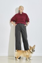 A woman dressed in a burgundy sweater and gray wide-leg pants stands confidently next to her corgi.