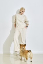 A woman dressed in light, casual attire stands against a plain wall while a corgi dog curiously