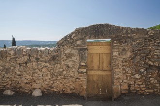Stone wall with wooden gate and view of a wide landscape under a blue sky, Provence, France