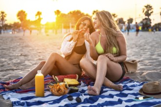 Two happy women sharing watermelon slices during a vibrant sunset picnic on the beach, enjoying