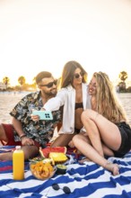 Group of friends taking a selfie during a sunset picnic on the beach, enjoying snacks and drinks