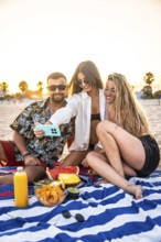 Three friends are taking a selfie with smartphone during a picnic at the beach, enjoying snacks and