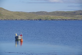 Lonely boat next to buoy on calm water, hilly landscape behind under clear sky, Hillswick,