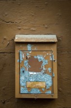 A rustic mailbox is mounted on a weathered brown wall, featuring peeling paint and faded colors.