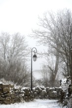 A vintage lamp post stands still amidst a snowy landscape, surrounded by frosty trees and a stone