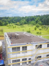 Building shell with crane and workers, surrounded by trees and clear sky, green roof, house