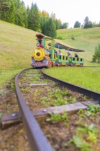 A colourful mini train moves through a green hilly landscape on narrow tracks, Retro Freizeitpark