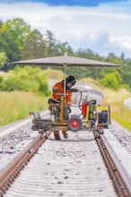 Worker using a machine on railway tracks under an umbrella, surrounded by greenery, grinding work