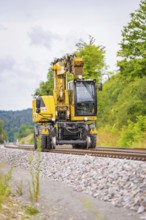 Yellow railway crane on railway tracks, surrounded by trees and ballast, grinding work on the