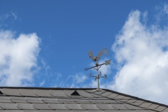 A metal weathercock on the roof in front of a blue sky with clouds