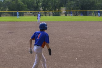 Young baseball player is walking on the infield during a game, wearing a blue and orange jersey