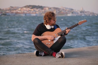 Hipster street musician in black playing electric guitar in the street on sunset on embankment with
