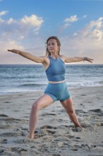 A woman performs Virabhadrasana 2 Warrior 2 pose on the beach, embracing the calm sunset and ocean