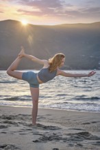 A woman performs Natarajasana, or Dancer's Pose, on a beach during sunset. The sun sets behind