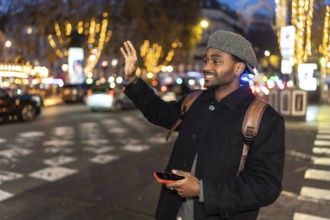 Young man smiling while hailing a taxi on a vibrant city street at night, holding a smartphone and
