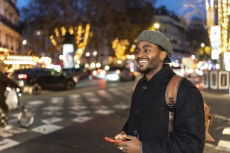 Smiling young black man wearing a winter coat and beret, carrying a backpack, using a phone while